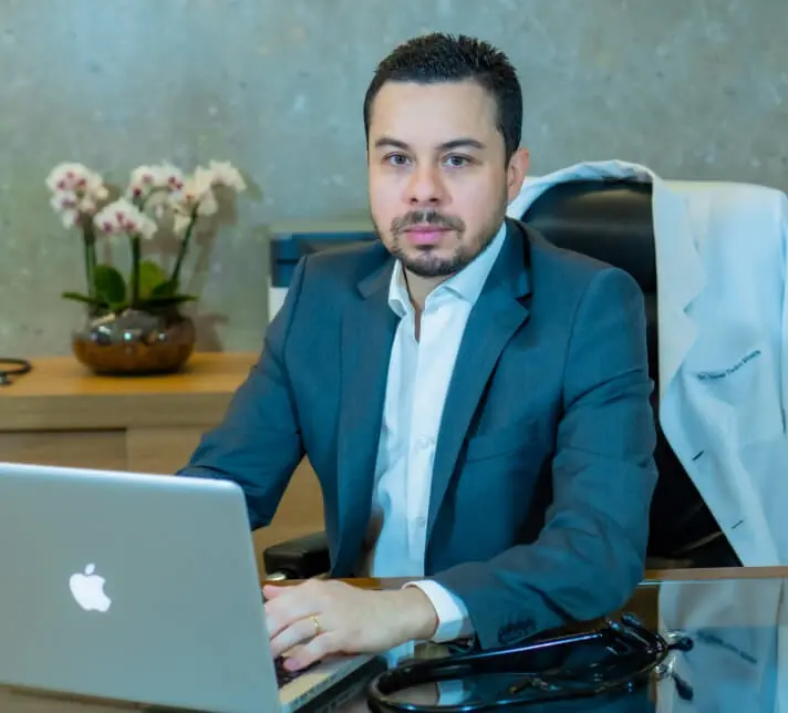 Dr. Lucas Tadeu Moura sentado à mesa do consultório, com notebook e estetoscópio, olhando para a câmera